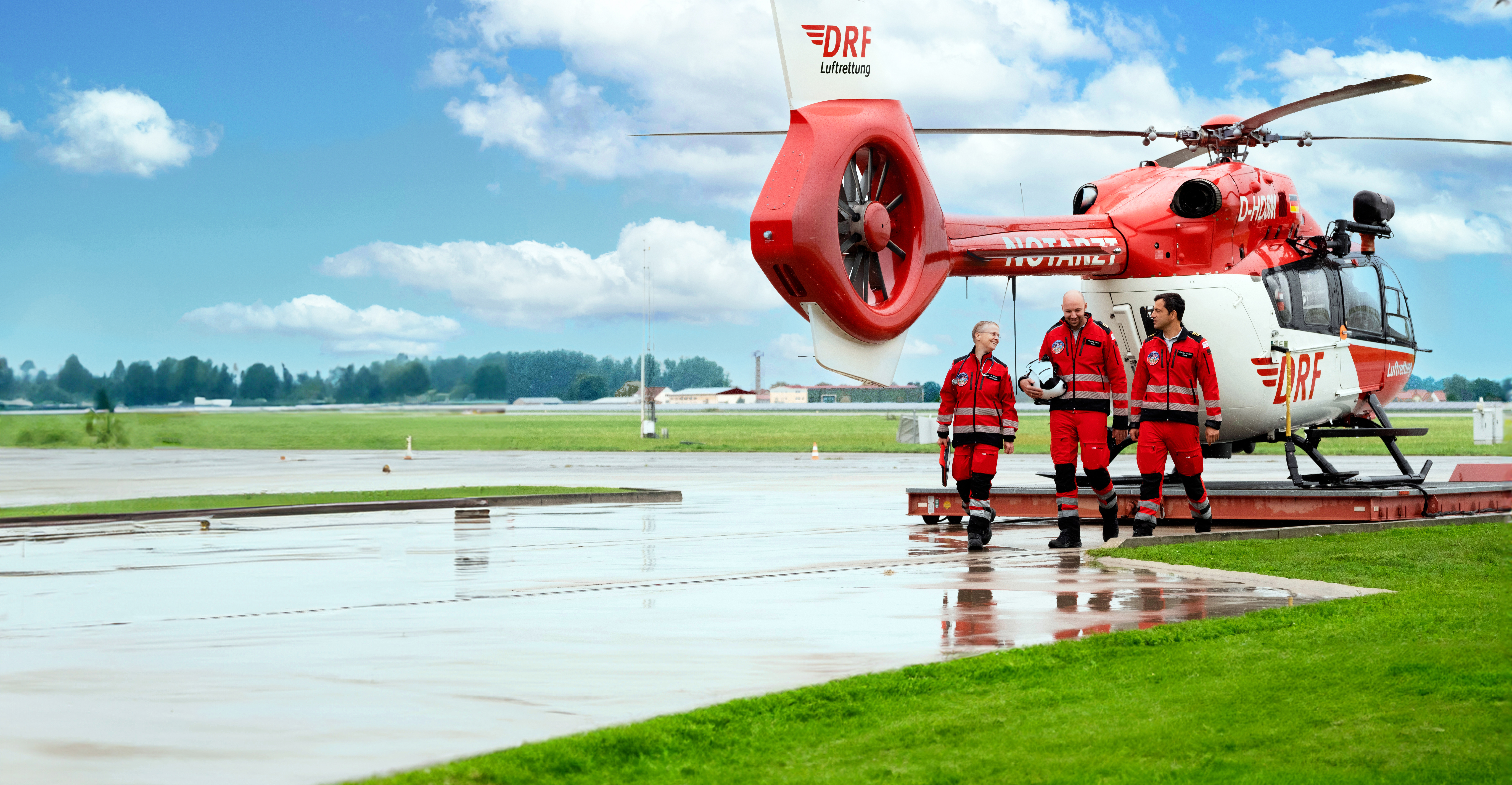 Die Crew eines Rettungshubschraubers läuft nebeneinander. Im Hintergrund ein Hubschrauber der DRF Luftrettung.